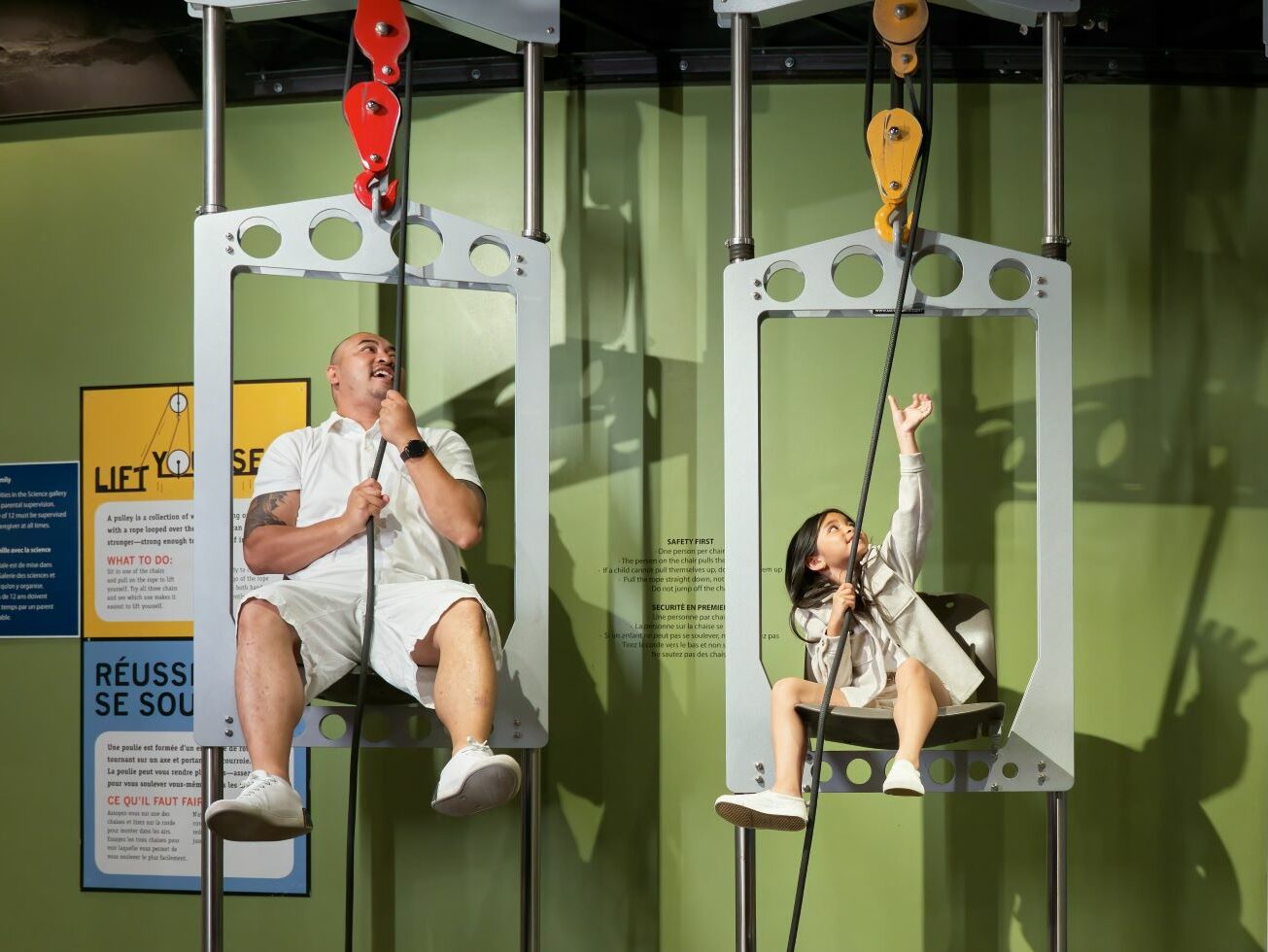 An adult and child race on the Pulley Chairs in the Manitoba Museum Science Gallery.