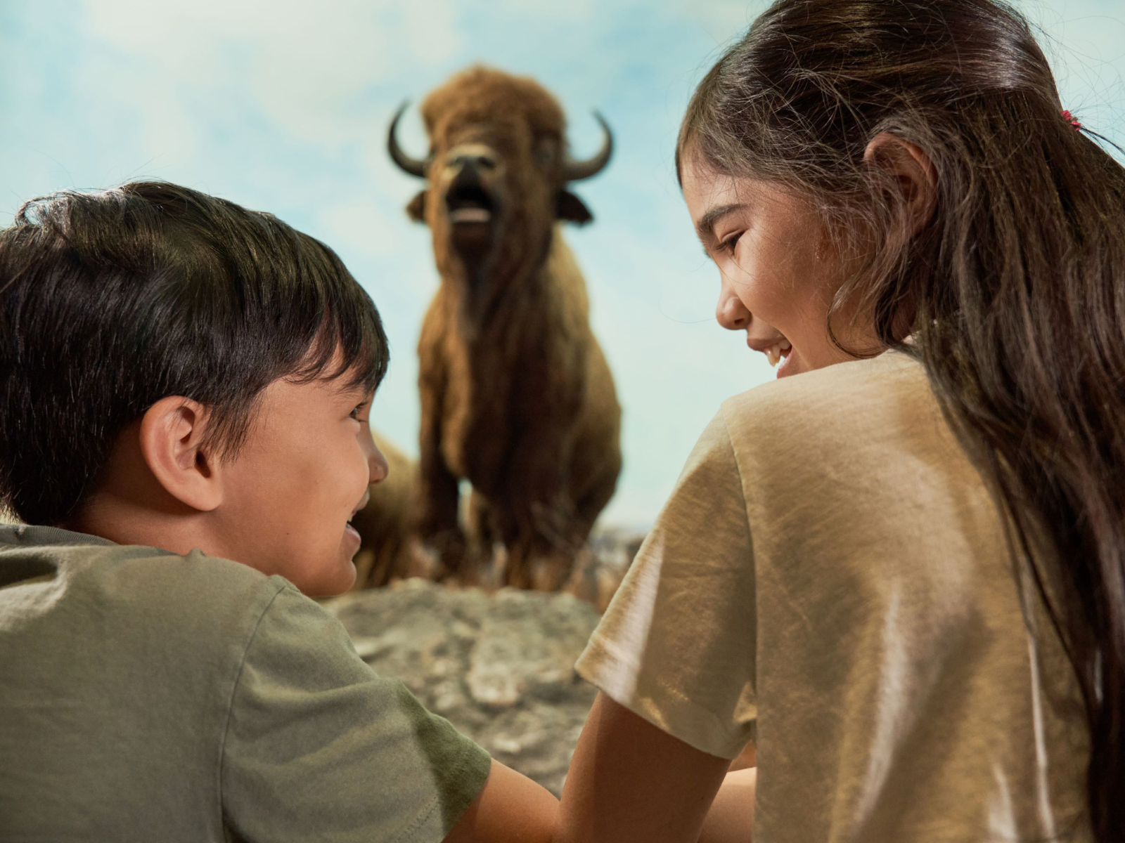 Two children smile at each other standing in front of a full-sized bison diorama.
