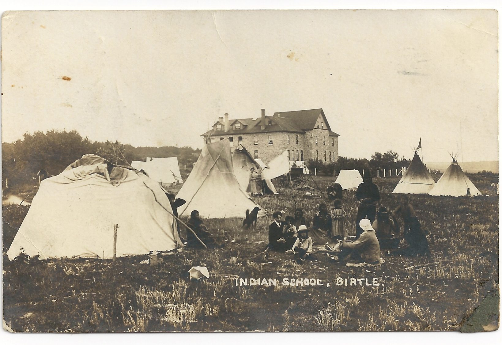 Sepia photograph. A group sits around a campfire outside of a Tipi. Several other Tipis dot the ground in front of a multi-storey building.