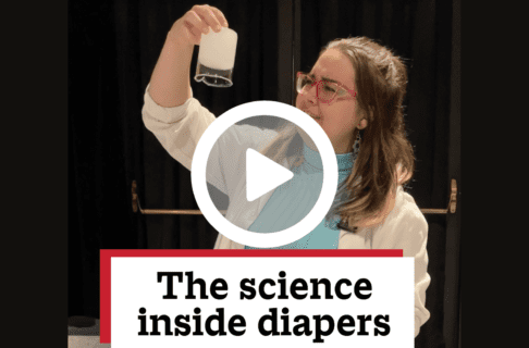 A screenshot of a video, an individual wearing a lab coat stands hold up an upside down full beaker in the Science Gallery. There's a play button over the screenshot and overlaid text reads, "The science inside diapers".