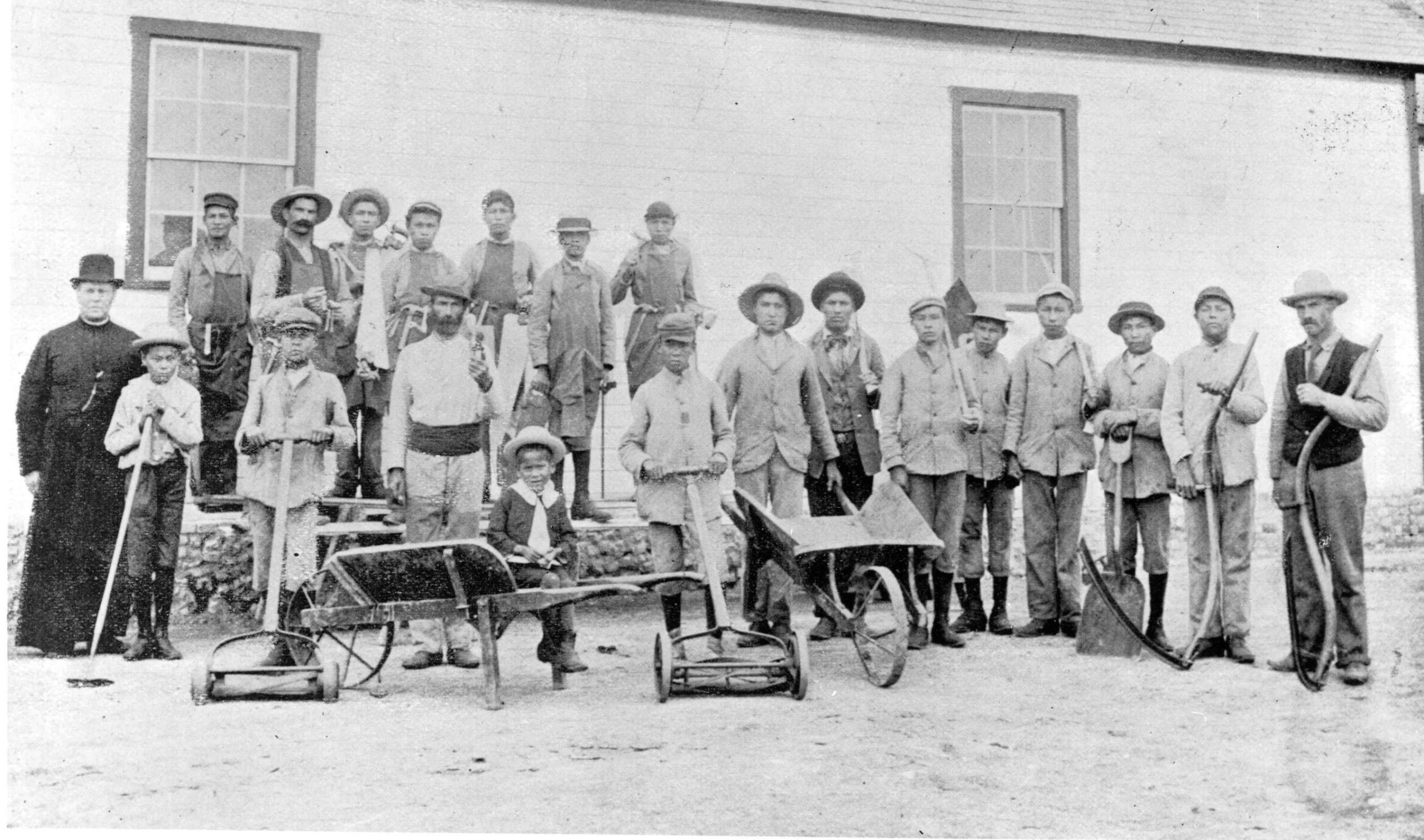 Black and white photograph of a couple dozen young people and adults standing in a group. Most are holding various hand tools, a few stand behind wheelbarrows or push-mowers.
