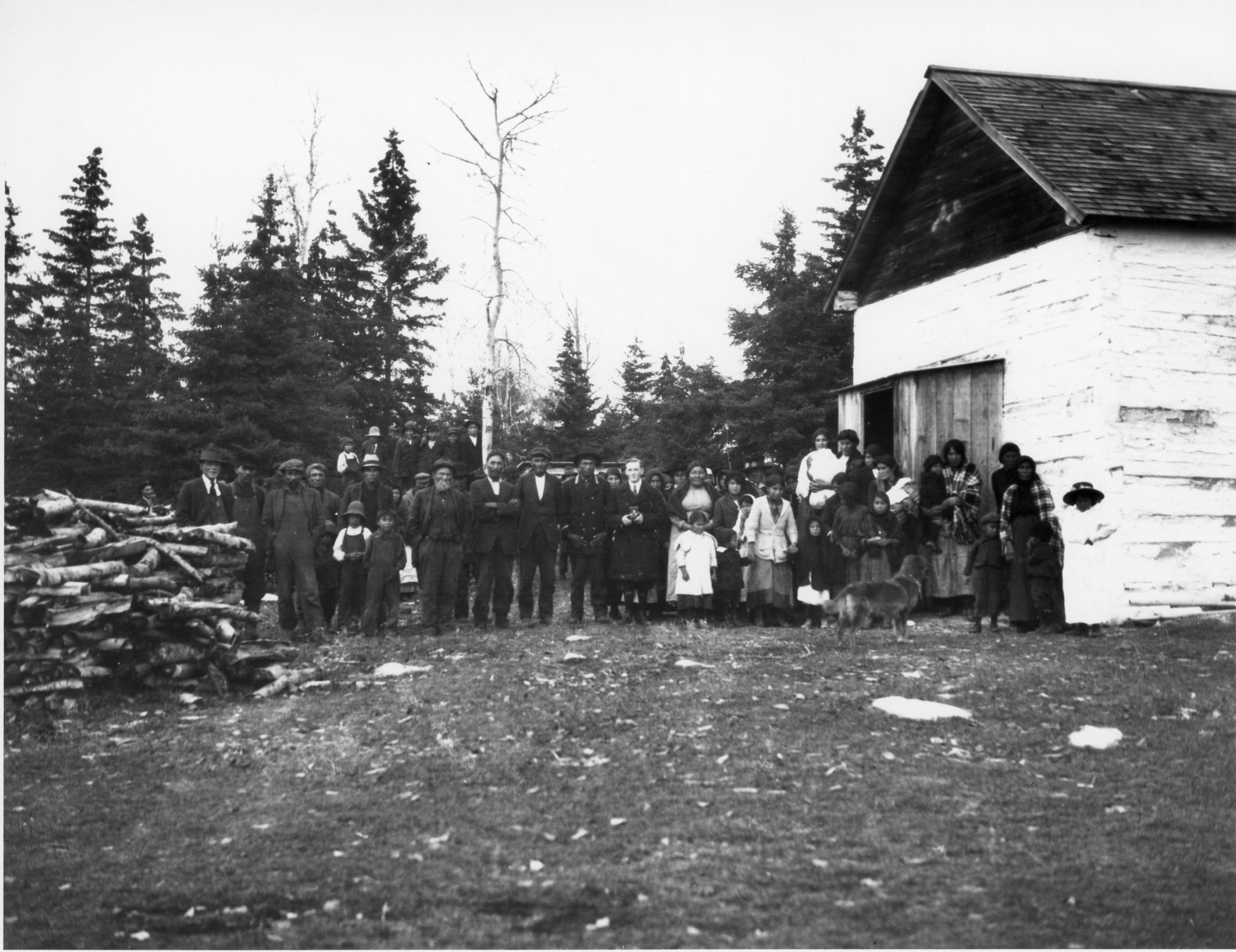 A group of several dozen people standing together outside in front of a treed area next to a single-storey wooden building.