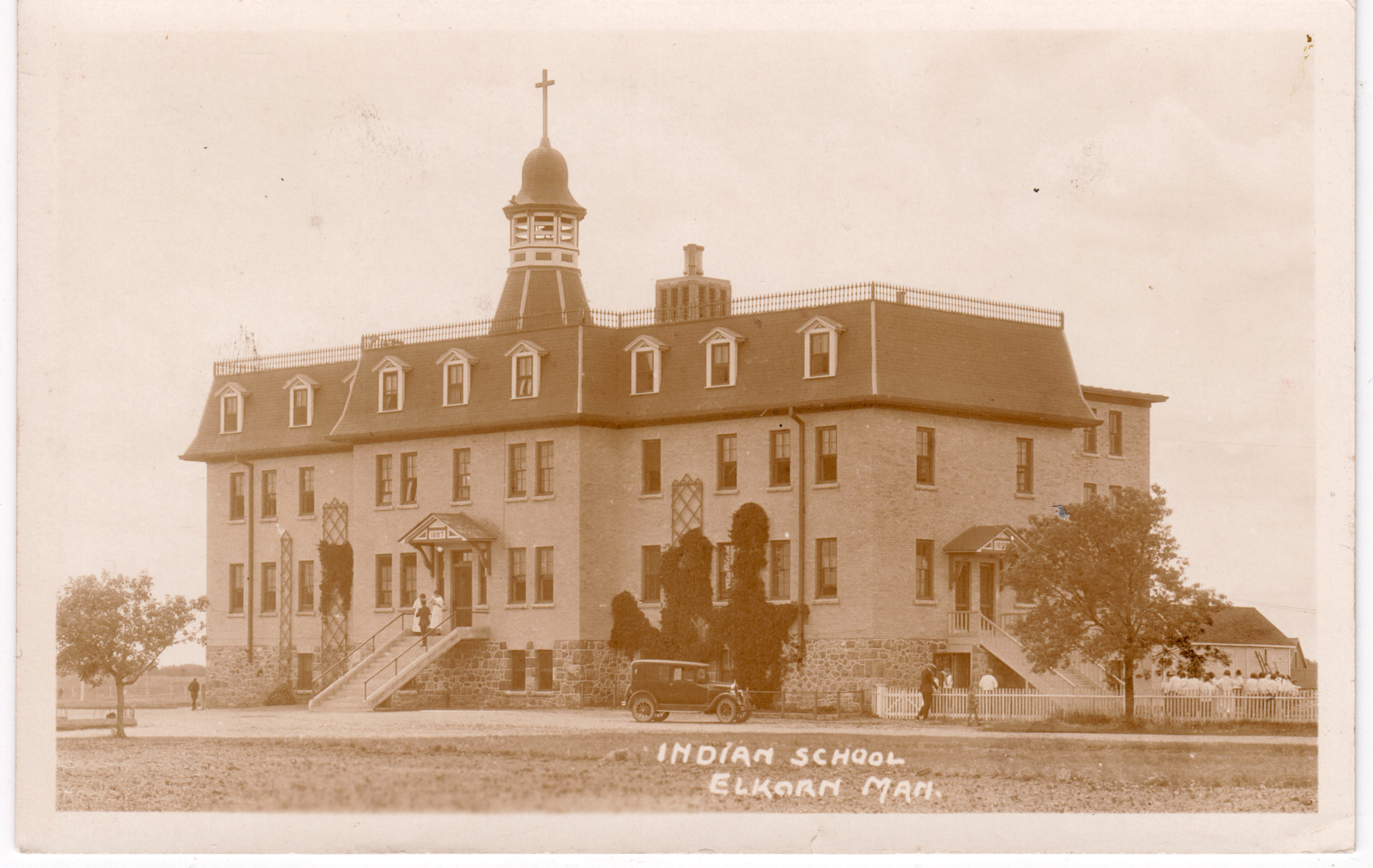 Sepia photograph of a three-storey building with a steeple bearing a cross. A vintage car is parked in front. Handwriting on the photo reads, "Indian School / Elkhorn Man".