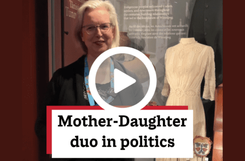 A screenshot of a video with an individual speaking to the camera standing in front of a Museum display case. There's a play button over the screenshot and overlaid text reads, "Mother-Daughter duo in politics".
