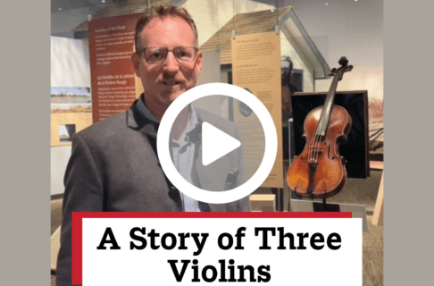 A screenshot of a video, an individual stands in front of a glass display cases containing a violin in the Museum Galleries. There's a play button over the screenshot and overlaid text reads, "A Story of Three Violins".