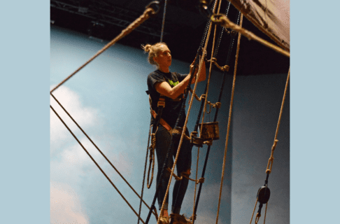 Woman wearing a pink harness and holding a paint brush with tar, on the Nonsuch rigging.