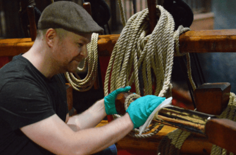 An individual wearing a flat cap and rubber gloves polishing a brass surface on a large wooden ship.