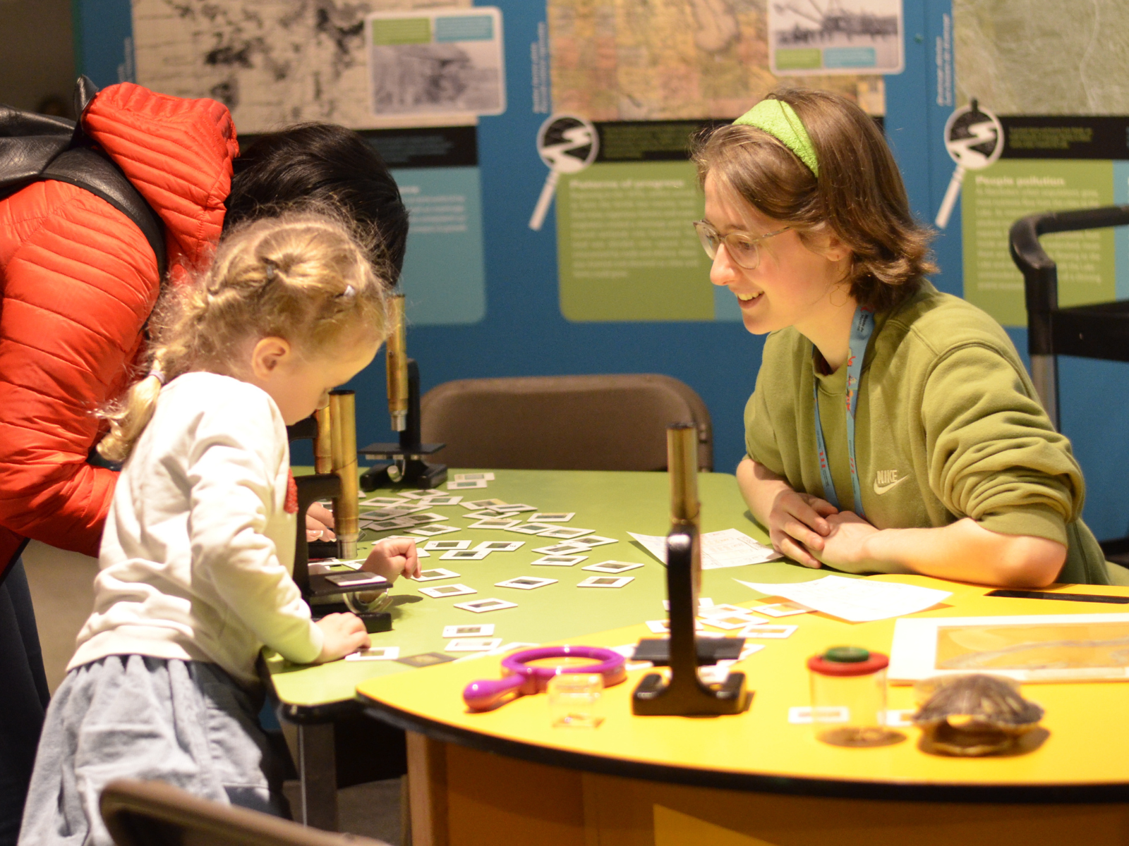 A Museum staff member seated at a table supervising a young visitor who is looking into a microscope.