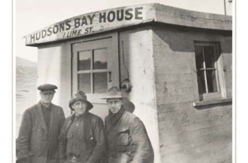 Black and white photograph of three individuals wearing coats and hats standing outside a small wooden building. On the overhanging eaves above their heads is painted “Hudson’s Bay House / 1 Lime St”.