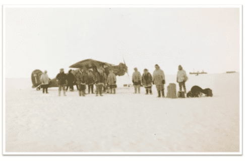 Sephia-toned photograph of a group of pepole in winter-wear standing on snowy ground beside a small airplane.