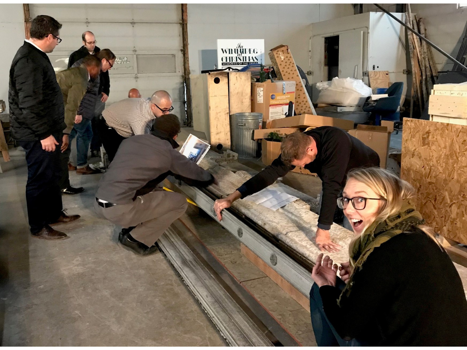 Several individuals standing and crouching around the pieces of the Eaton’s lintel and metal support beam in a storage space. Conservator Carolyn Sirett is turned, facing the camera, grinning.