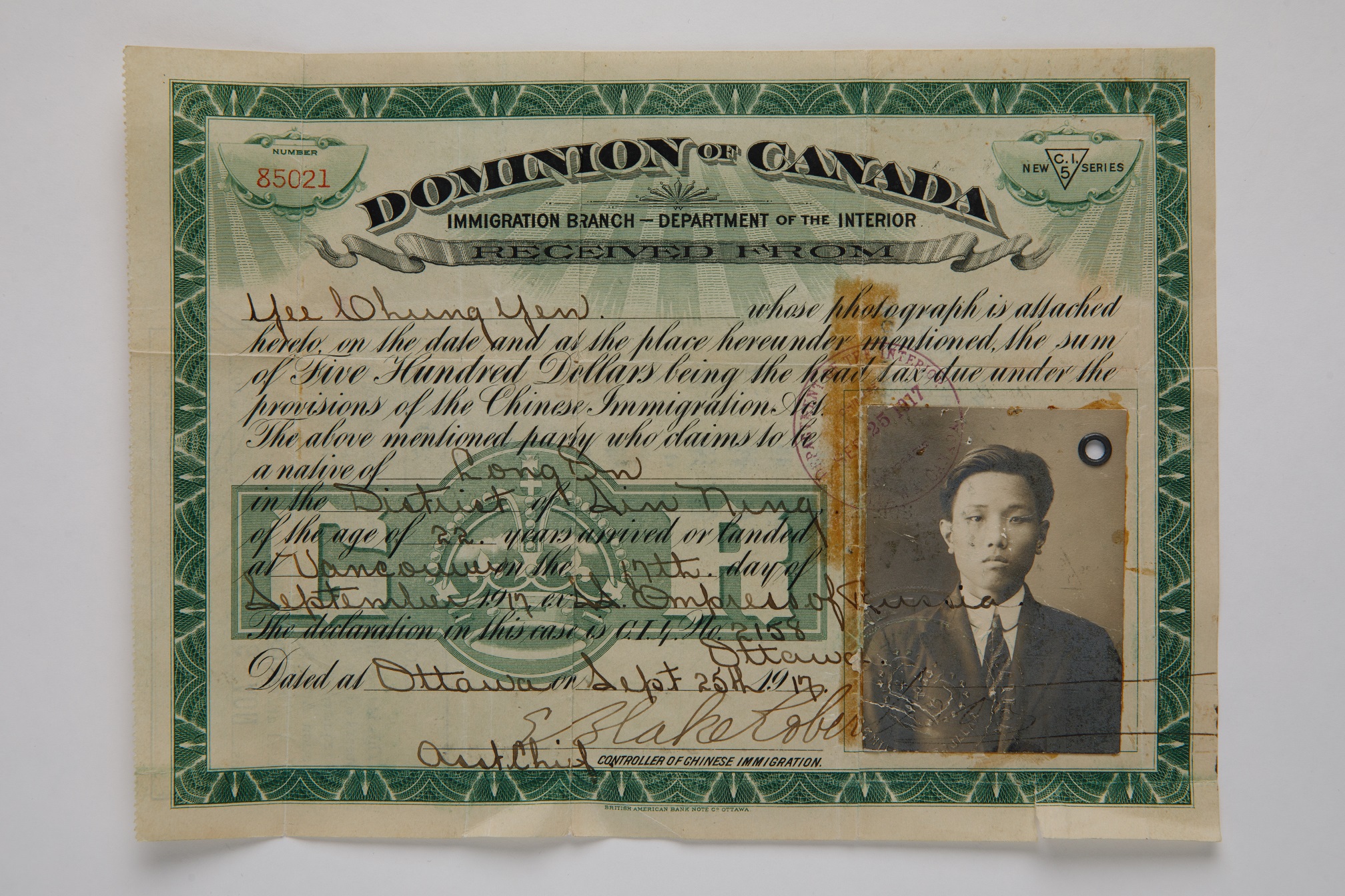 Photograph of a Head Tax Certificate. Along the top reads, “Dominion of Canada / Immigration Branch – Department of the Interior”, and in the bottom right corner is a identifiaction photo of a serious-looking young man wearing a dark suit.