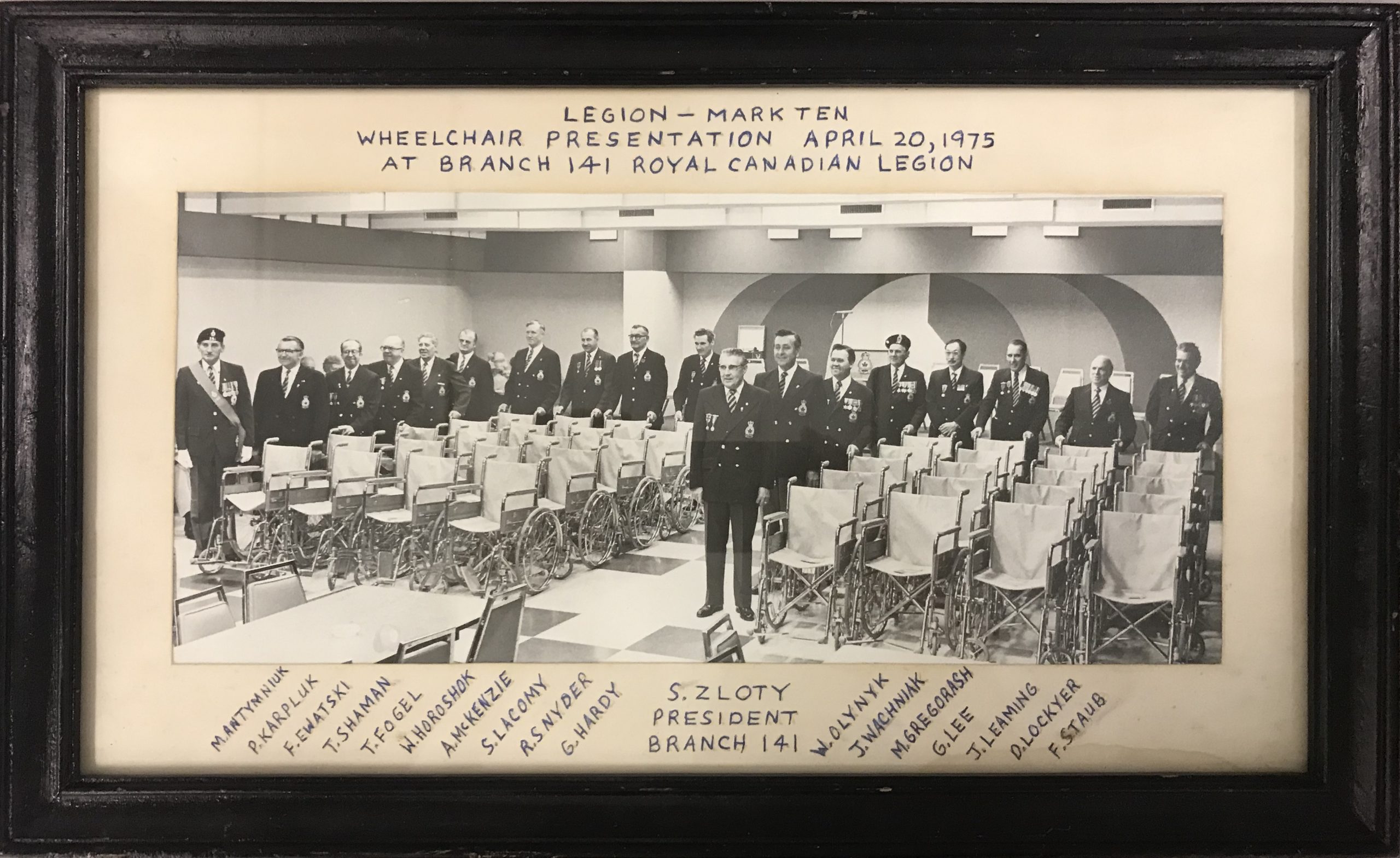 A framed black and white photograph showing a group of uniformed men standing alongside neatly lined up empty wheelchairs. Written on the frame surrounding the photo, writing reads, “Legion - Mark Ten / Wheelchair Presentation April 20, 1975 at Branch 141 Royal Canadian Legion.” Written along the bottoms are the names of those pictured including S. Zloty, President Branch 141.