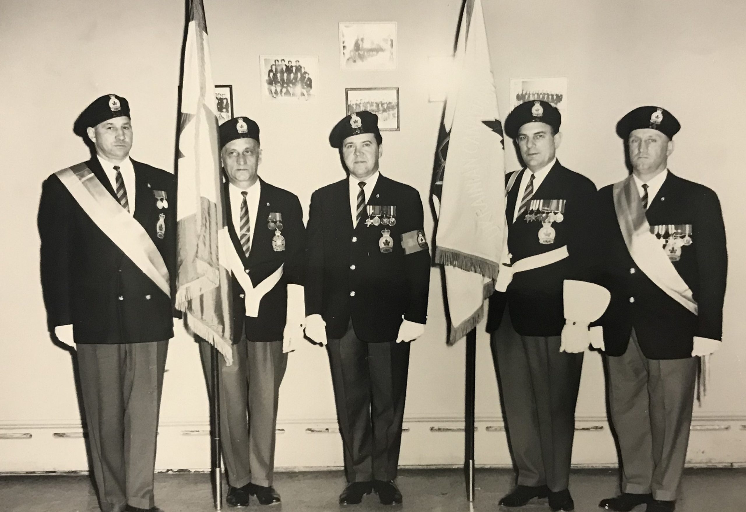 Black and white photograph of five men in uniform standing on a row with serious faces. Two of them are holding up flags draped around their poles.