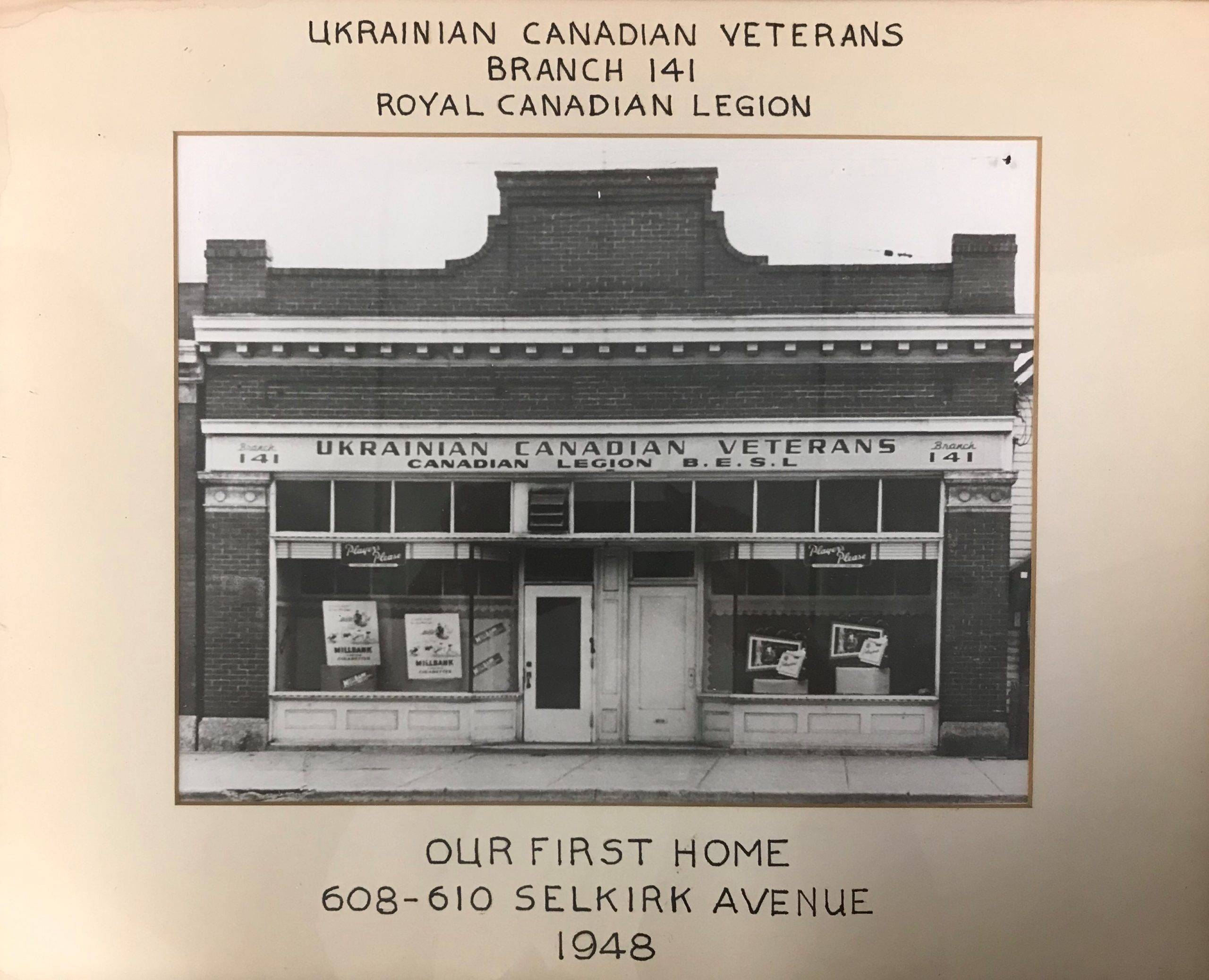 A framed black and white photograph of a brick building exterior with a sign reading, “Ukrainian Canadian Veterans / Canadian Legion B.E.S.L.”. Written on the frame surrounding the photo, writing reads, “Ukrainian Canadian Veterans / Branch 141 / Royal Canadian Legion / Our First Home / 608-610 Selkirk Avenue / 1948”.