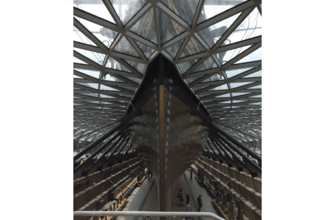 View of a large ship’s keel displayed in dry dock hall where viewers can get up close with is. A glass ceiling has been built at “water level” through which one can see the rest of the ship above.
