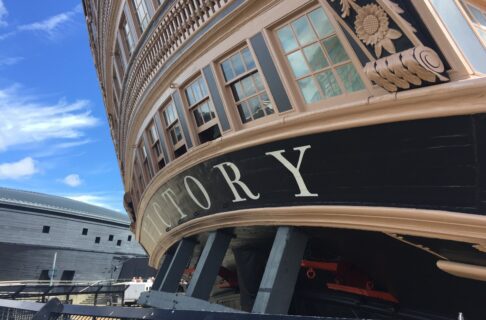 A close-up of the stern of a large wooden ship with “VICTORY” written across the back. In the background is a domed black building.