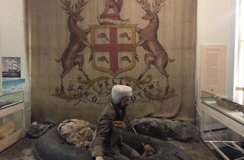 A mannequin wearing a jacket and light-coloured fur hat sitting in a small boat with a pack. Behind the mannequin is a large wall handing with the HBC Coat of Arms. The coat of arms bears two rearing elk either side of a shield with a beaver in each of the four sections of a cross. A fox sits above the shield, and a banner below reads, “Pro Pelle Cutem”.