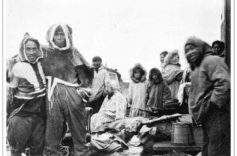 Black and white photograph of a group of individuals in winter-wears beside a piles with some supplies and barrels. A few of the group are looking and smiling at the camera.