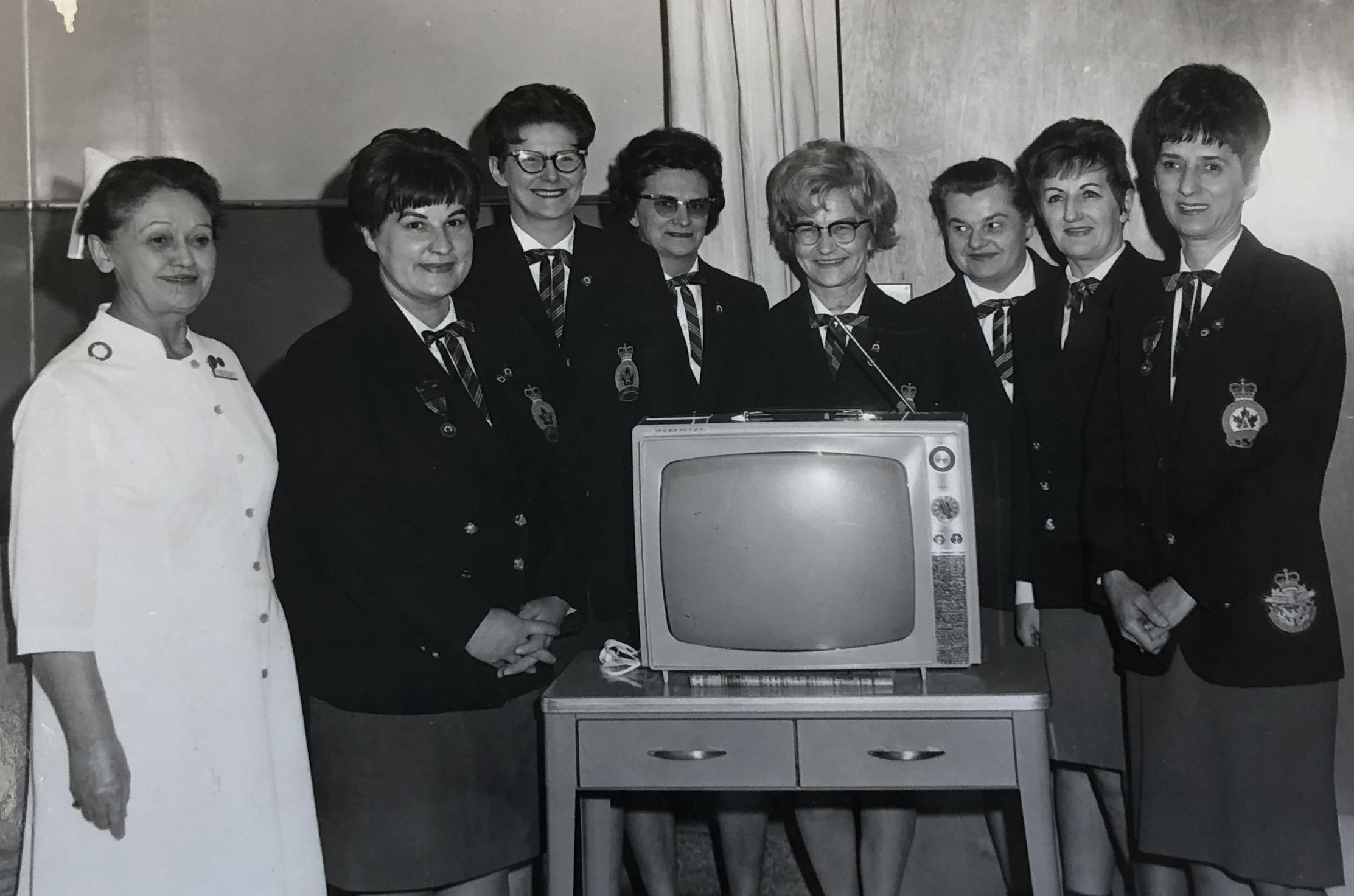 A black and white photograph showing seven women in dark Women’s Auxillary uniforms standing beside a woman in a white nurse’s uniform around a vintage television set placed on a small desk.