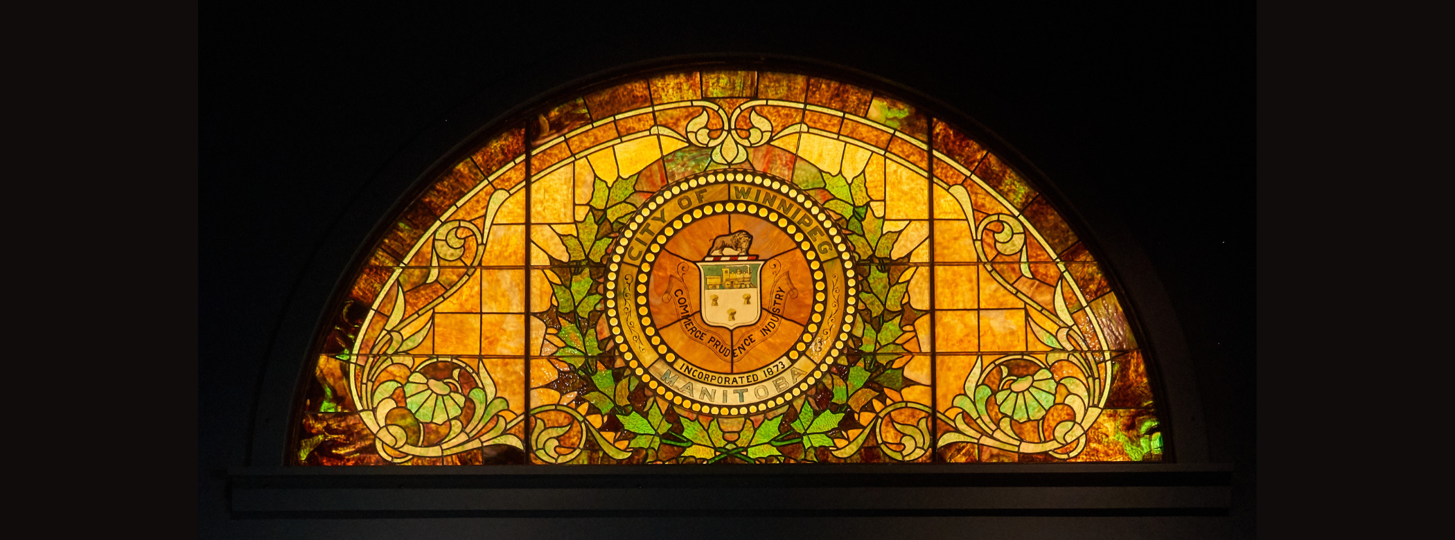 Large half-circle stained glass window installed on a dark wall, allowing the lit glass to stand out in contrast. The stained glass features a City of Winnipeg crest in the middle framed by a green wreath.