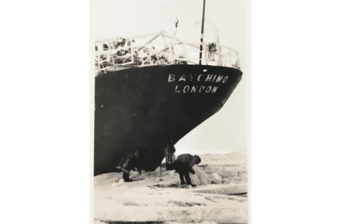 Black and white photograph of two individuals on thick ice below the stern of the ship “BAYCHIMO”.