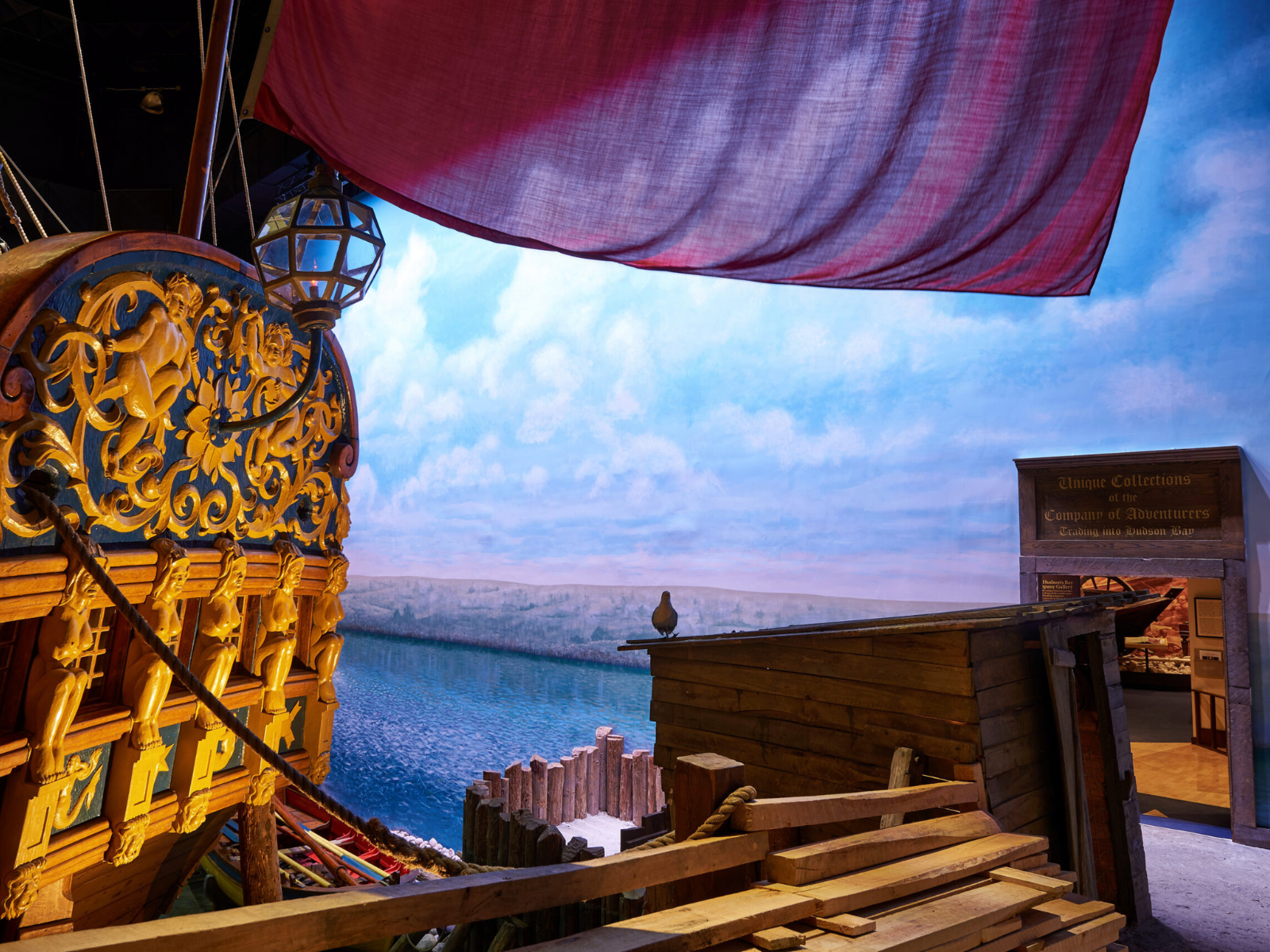 Photograph looking past the stern of a large wooden sip (the Nonsuch) toward a painted background mural showing blue water and a hilly coastline in the distance.