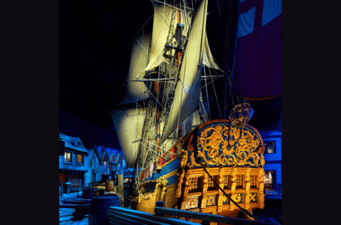 Looking toward a wooden sailing ship (the Nonsuch) from the stern as it is lit dramatically within a blue-hued gallery. The ship is displayed docked in part.