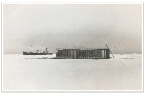 Black and white photograph of a single story building on flat snowy ground. In the distance a steam ship can be seen.