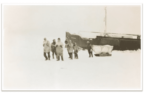 Black and white photograph of a small group pulling a sled with what appears to be a small boat on it as they move away from a larger ship in the ice.