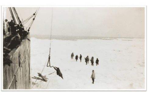 Black and white photograph from the deck of a tall ship looking down onto the ice below where a small group stands watching as supplies are lowered on a hook.