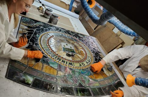 Conservator Carolyn Sirett (left) and Conservation Technician Loren Rudisuela (right) working either side of a work bench, putting putty onto the new lead came of a large stained glass window.