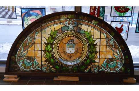 Large half-circle stained glass window propped up on wooden supports in front of a window. The stained glass features a City of Winnipeg crest in the middle framed by a green wreath.