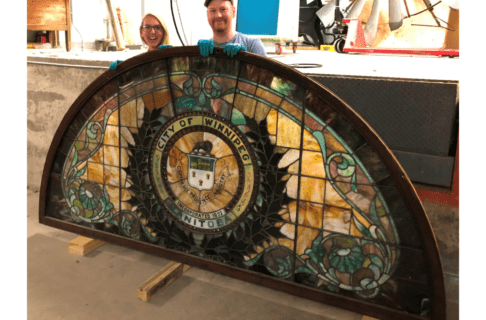 Conservator Carolyn Sirett and Conservation Technician Loren Rudisuela standing smiling at the camera behind a large half-circle stained glass window on wooden supports. The stained glass has a City of Winnipeg crest in the middle.