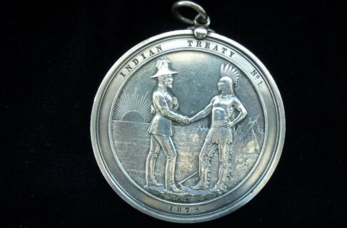 Photograph of a Treaty Number 1 handshake medal. A circular silver medal portraying a representitive of England shaking hands with a First Nations leader. They stand on grassy ground in front of tipis and the rising sun. Text around the edge of the medal reads, “Indian Treaty No. 1 / 1873”.