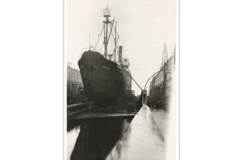 Black and white photograph of a tall steam ship in a narrow dock.