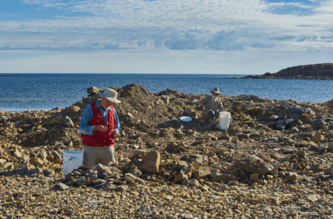 Dr. Graham Young, wearing a red vest with a white pail beside him, kneels on a rocky shoreline in front of a pile of rocks, examining two thin pieces more closely. Further back to the right side of the photo another individual sits on a rock with a pail in front of them. In the background is open water.