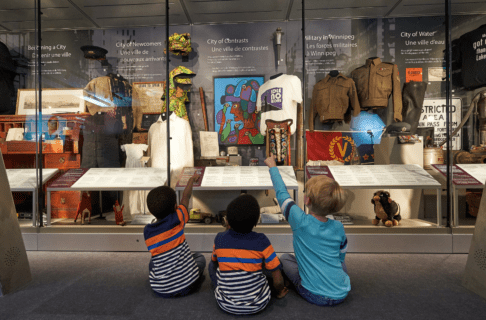 Three small children sitting on the carpet in front of a glass display case of various personal historical items, including army uniforms, traditional clothing, Indigenous art, and a stuffed animal.