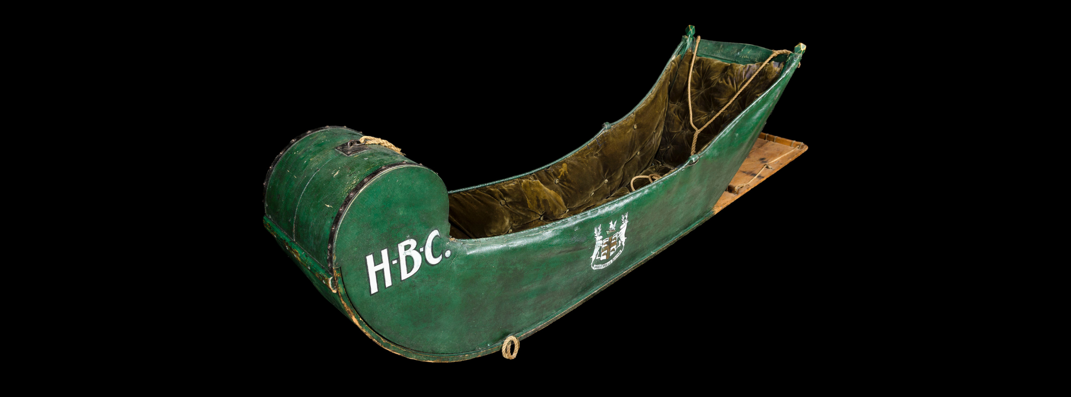 Sled (called a cariole) covered with green painted canvas and interior lined with plush olive-green velvet. The letters HBC are painted in white on the curved front, and the Company’s Coat-of-Arms painted on the side in white and red. The Coat-of-Arms features two elk flanking a crest with St. George’s cross in red and four little beavers in each quadrant, topped with a fox sitting on a cap, and a banner with their Latin motto Pro Pelle Cutem scrolled along the bottom.