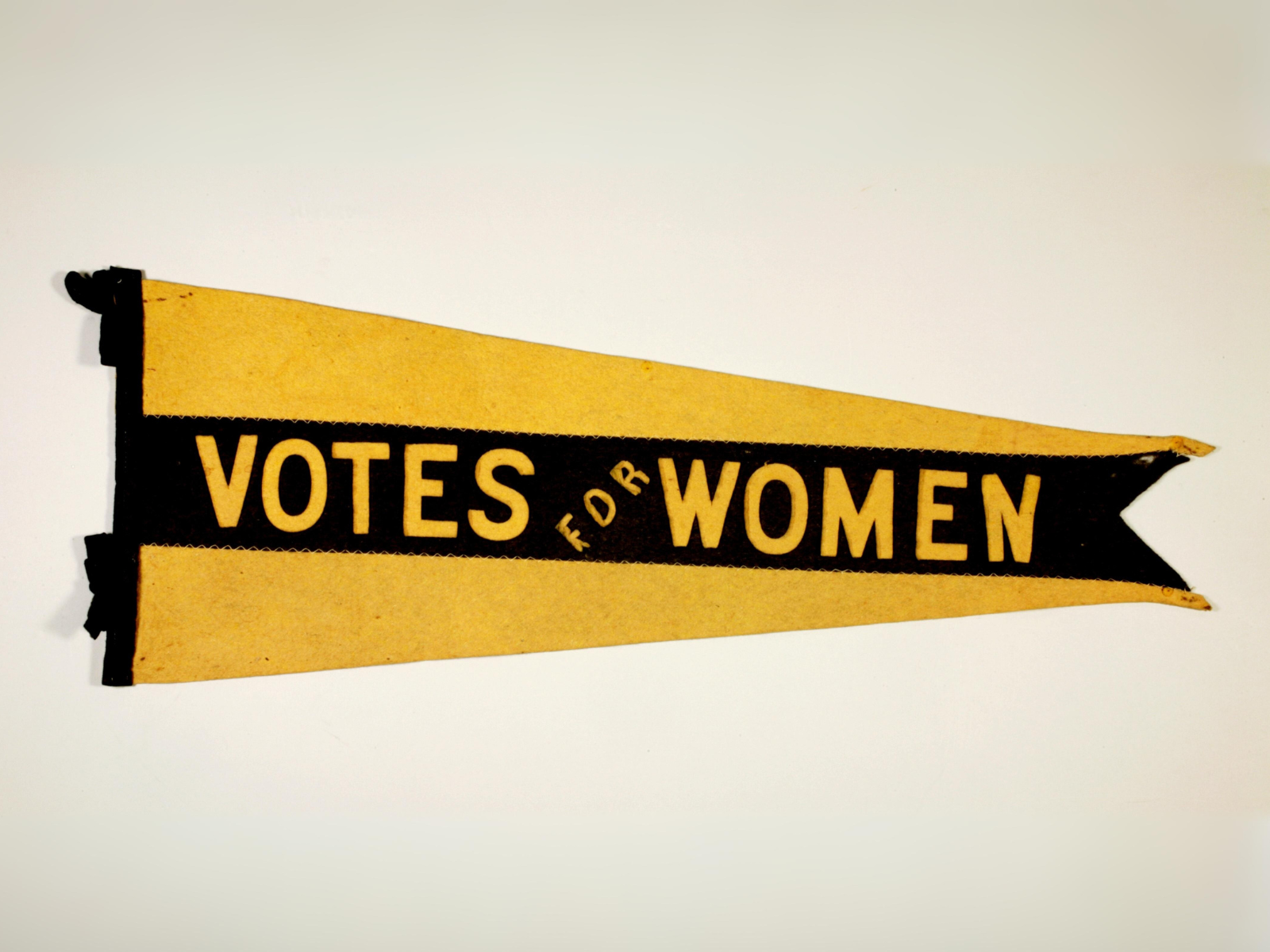 Elongated triangular flag. The background is yellow wiith a thick black stripe stitched on top reading “Votes for Women” along the centre.