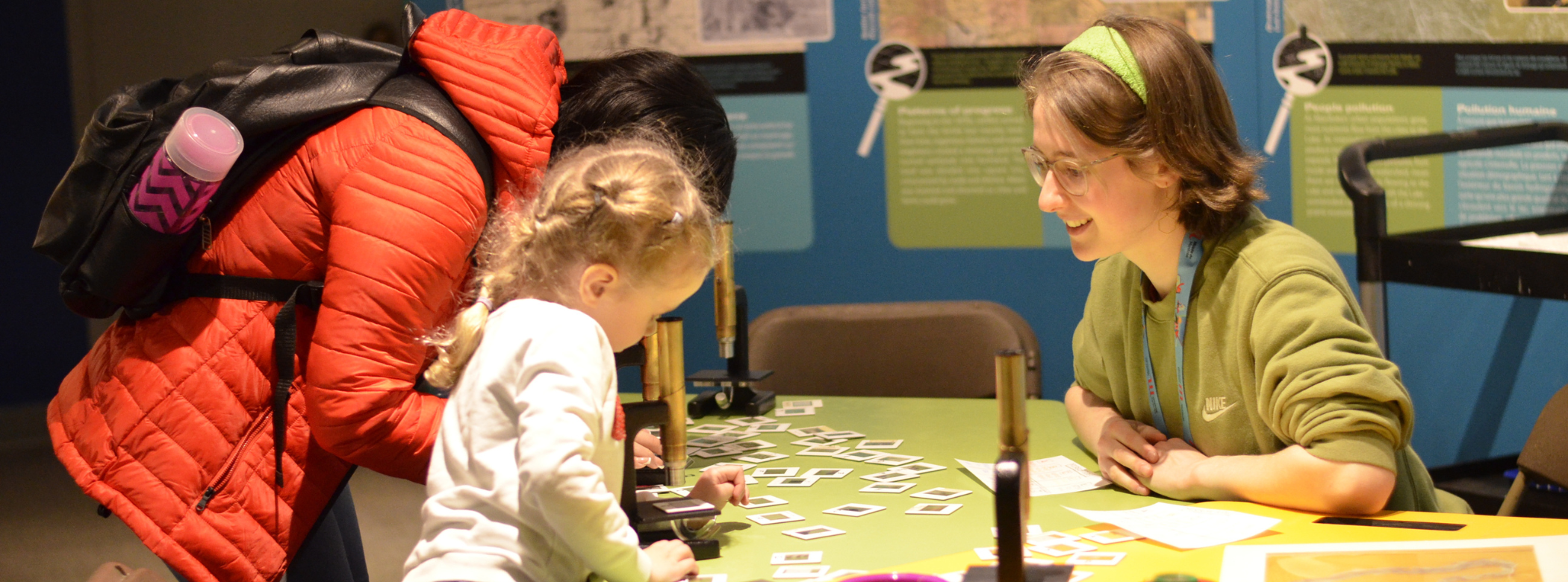 A Museum staff member seated at a table supervising a young visitor who is looking into a microscope.