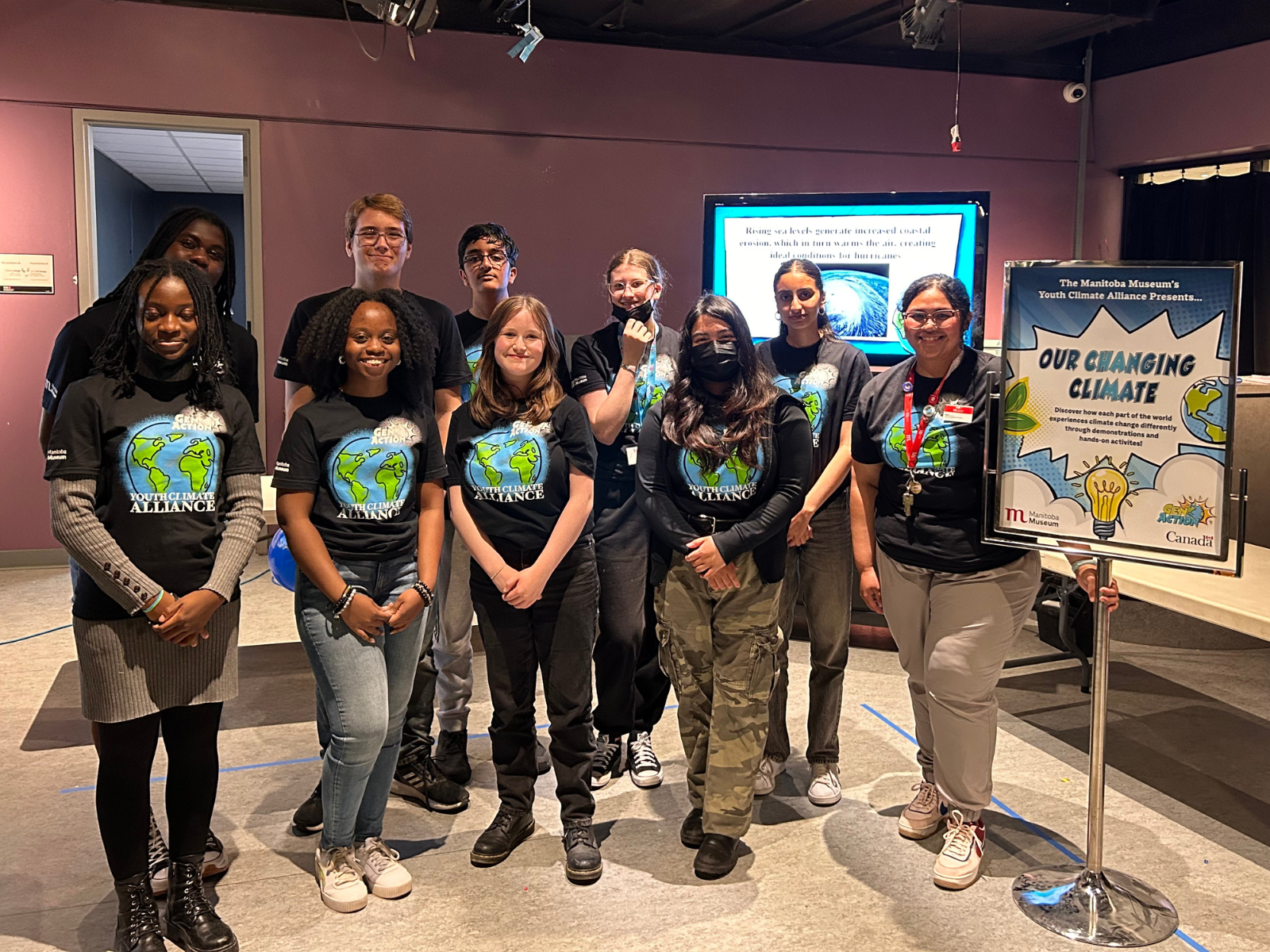 A group of nine youth and a Museum staff member smiling together. All are wearing matching t-shirts with an illustrated globe and the words “GenAction! / Youth Climate / Alliance”. On the right side of the group is a sign reading, “Our Changing Climate”.