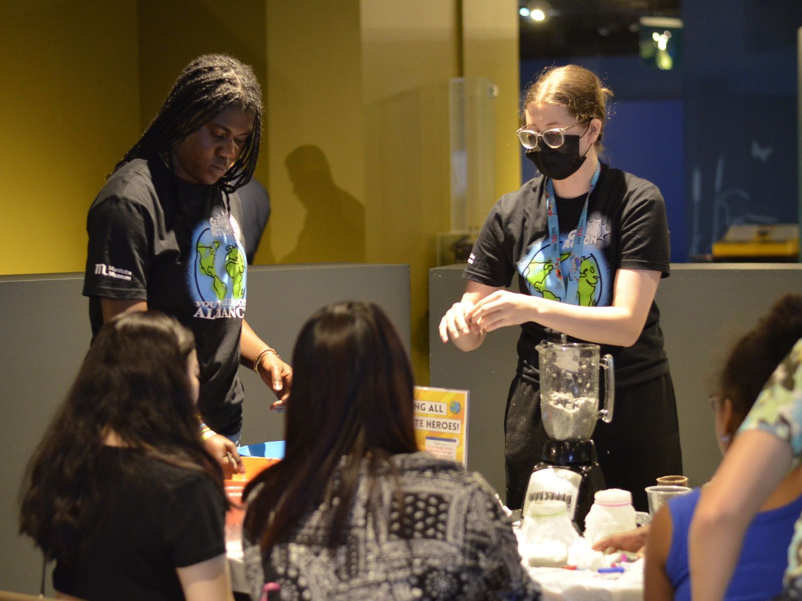Two youth running a pop-up exhibit in the Science Gallery for two visitors. Both youth are wearing matching t-shirts with an illustrated globe and the words “GenAction! / Youth Climate / Alliance” and a Manitoba Musuem logo on the sleeve.