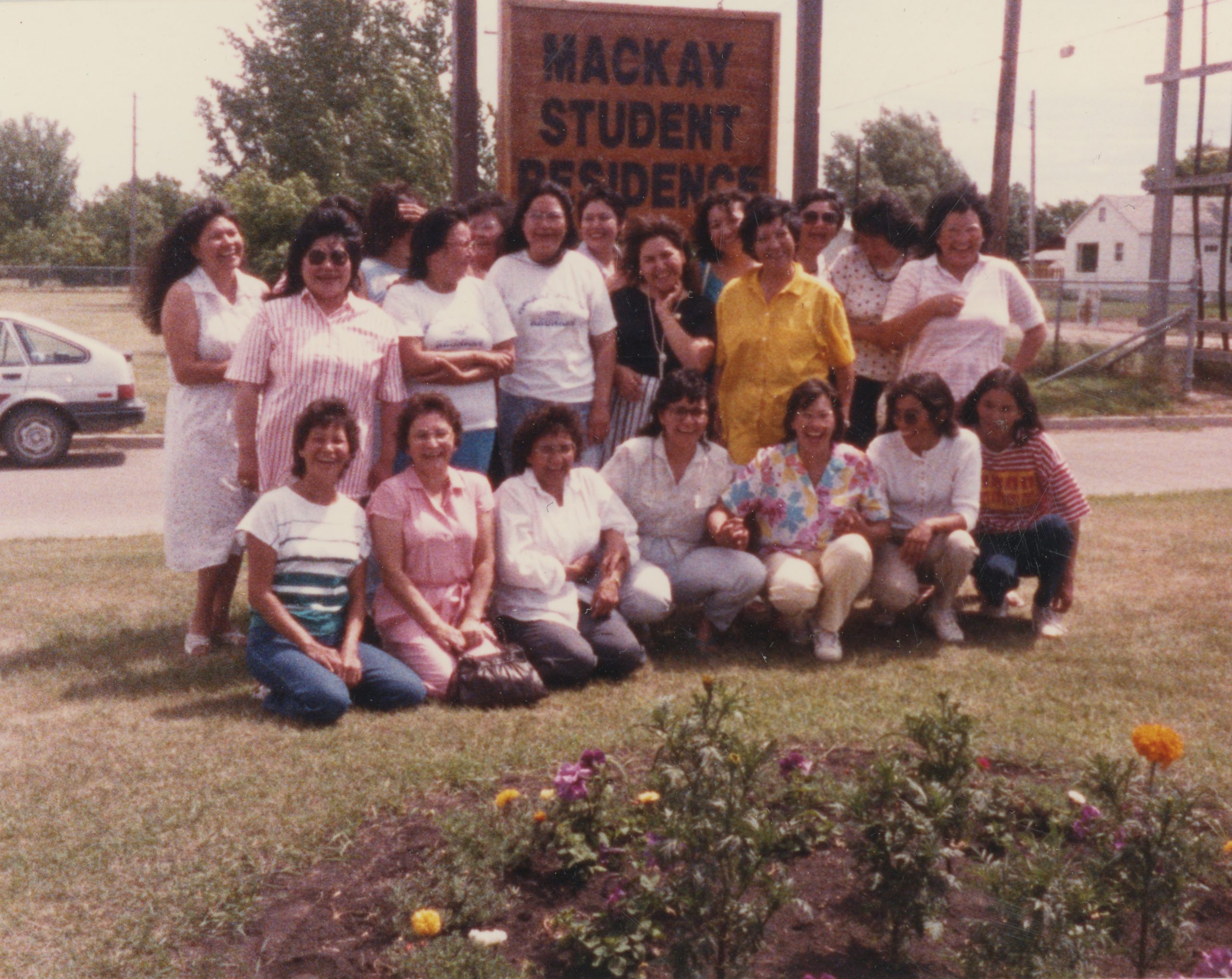 A group of twenty adults posing smiling in front of a MacKay Student Residence sign.