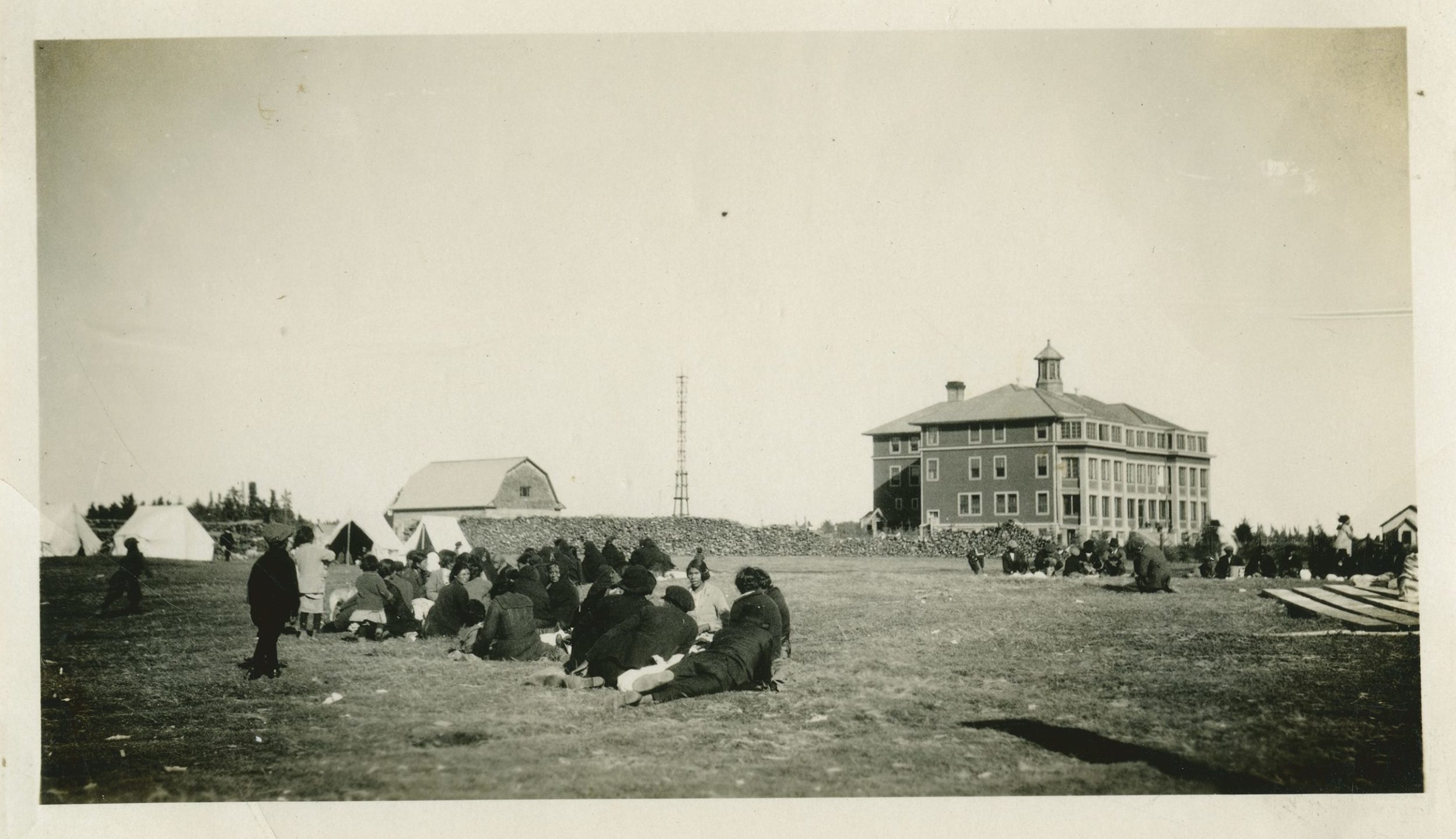 Sepia photograph of groups of people clustered on the flat ground in front of a three-storey building.