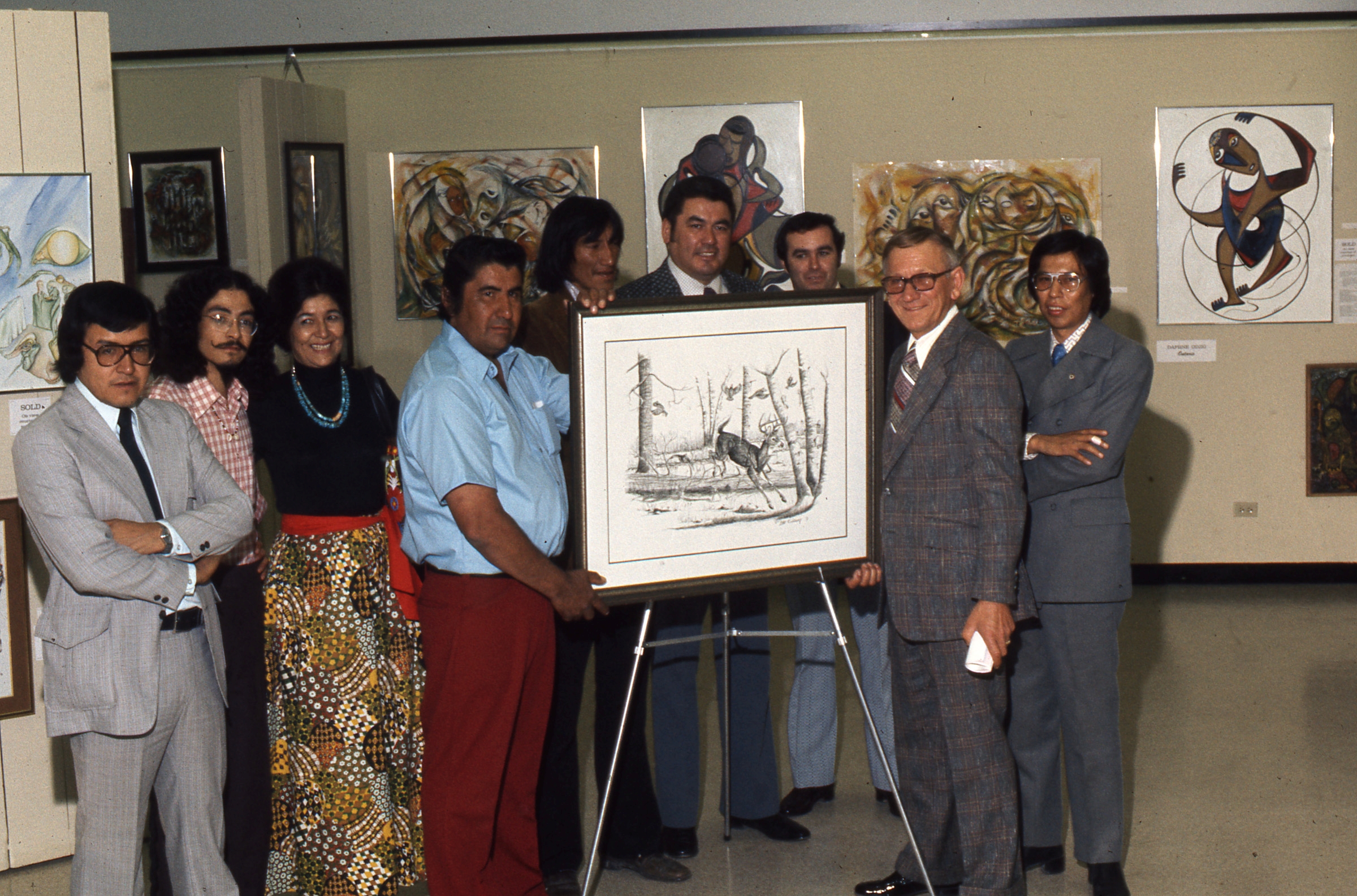 Nine people pose for a photograph beside a art piece on an easel. In the background, numerous artworks are mounted on the walls.