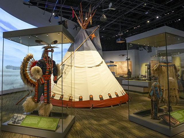 Photo of the Prairie Gallery at the museum with cases including regalia and a teepee in the background.