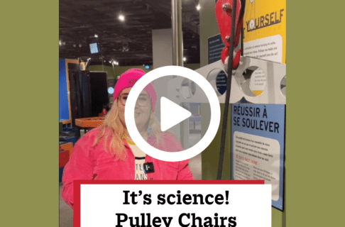 A screenshot of a video, an individual stands in front of a pulley chairs in the Science Gallery, speaking to the camera. There's a play button over the screenshot and overlaid text reads, "It's science! Pulley Chairs".