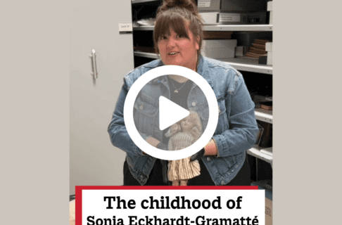A screenshot of a video, an individual standing in a collections space holding up a vintage doll, speaks to the camera. There's a play button over the screenshot and overlaid text reads, "The childhood of Sonia Eckhardt-Gramatté".