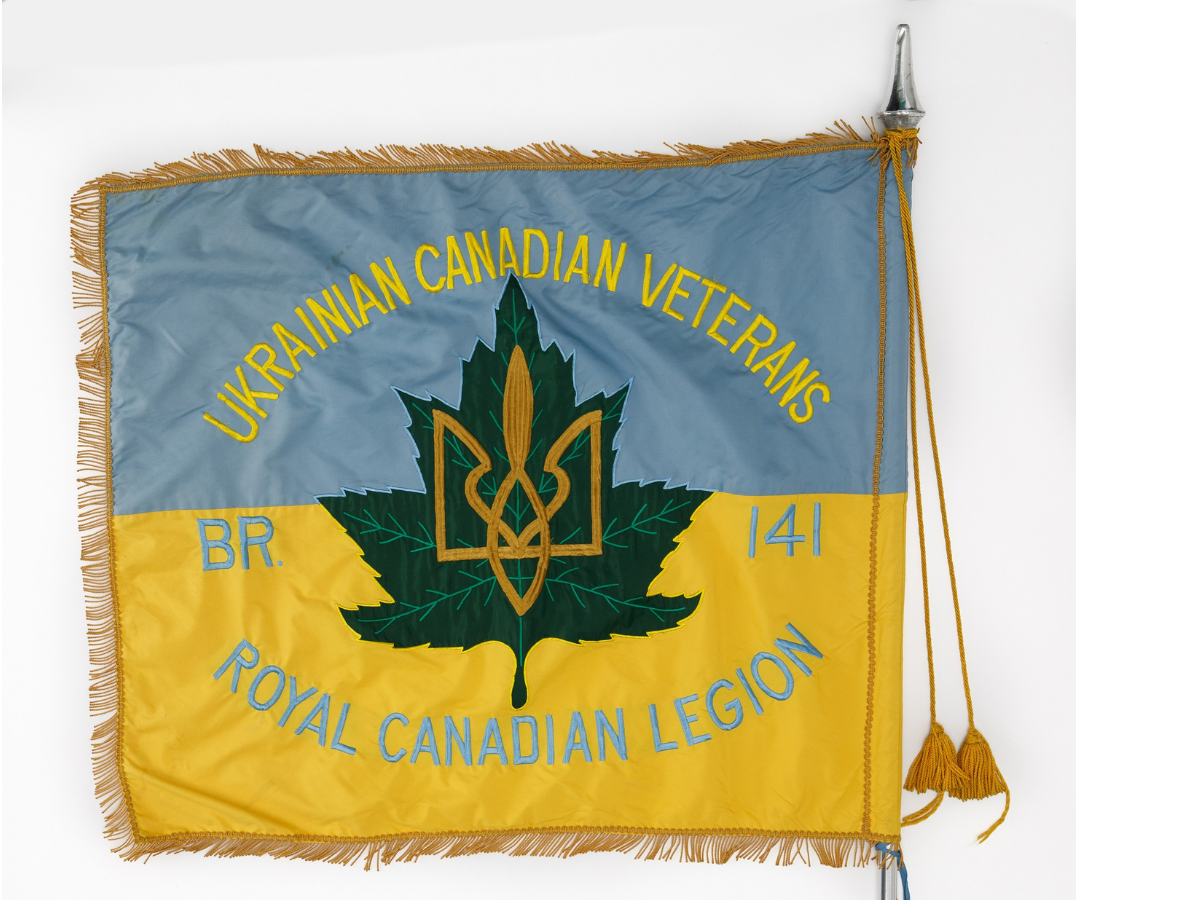 The Ukrainian Canadian Veteran’s flag. The top half of the flag is light blue, and the bottom half is yellow. In the centre is a dark green maple leaf with a gold symbol on it – the Tryzub, or Ukrainian “trident” symbol. In yellow thread on the upper half of the flag is stitched, “Ukrainian Canaidna Veterans”. In light blue thread on the lower half of the flag is stitched, “Br. 141 / Royal Canadian Legion”. The flag has a gold fringe around the edges.